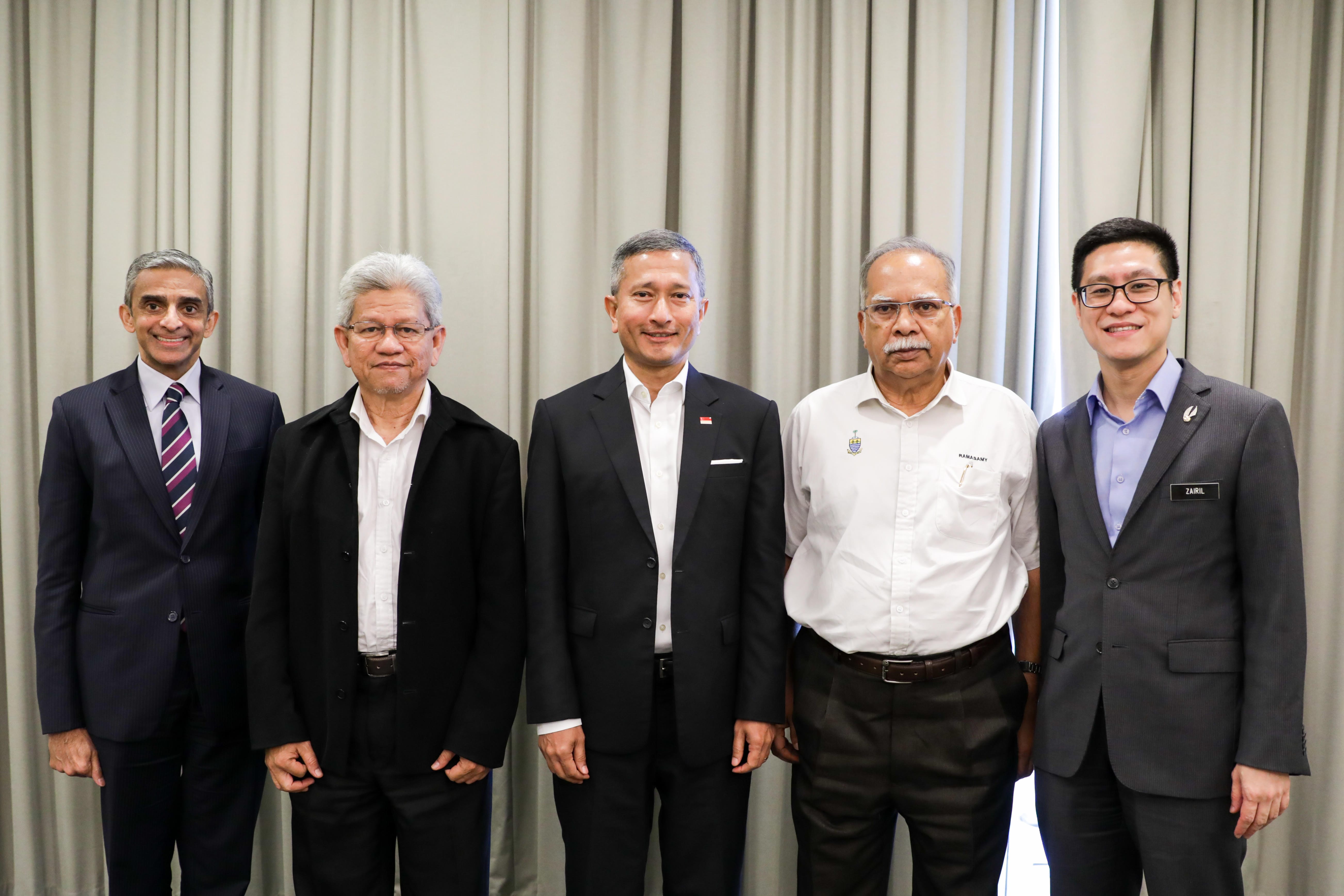 Five men stand in suits against a neutral curtain backdrop.